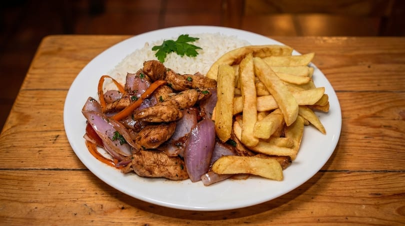 Lomo Saltado with chicken, red onions, tomatoes, french fries, and white rice on a wooden table.