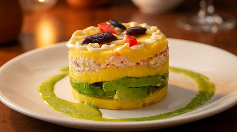 Peruvian causa, a layered dish of yellow potato, avocado, and crab, garnished with olives and red pepper.