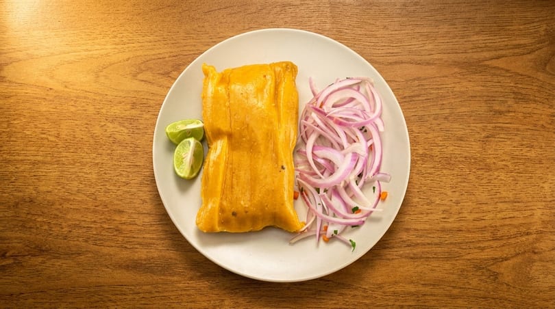 Peruvian tamale with red onion salad and lime wedges on a white plate, on a wooden table.
